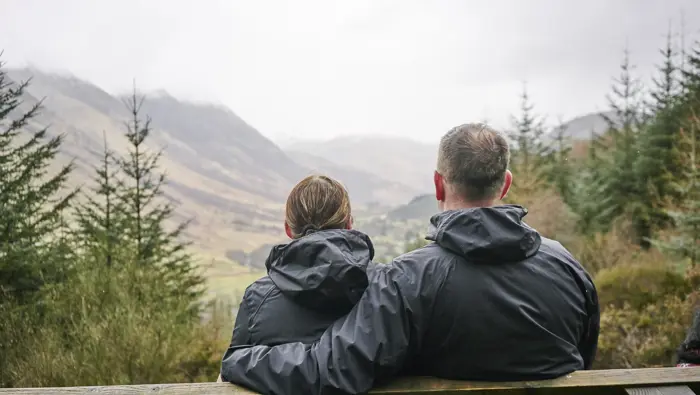  Man and woman both in blue waterproof jackets sit on wooden bench admiring view over woodland and valley towards a mist covered and rainy Ben Nevis and adjoining mountain range, at Glen Nevis