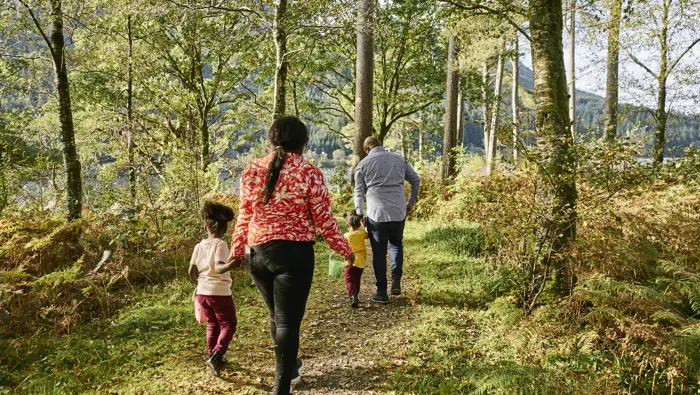 Rear view of man and woman walking along tree lined loch side path with two little girls carrying plastic bucket and spade, Jubilee Point, Loch Eck