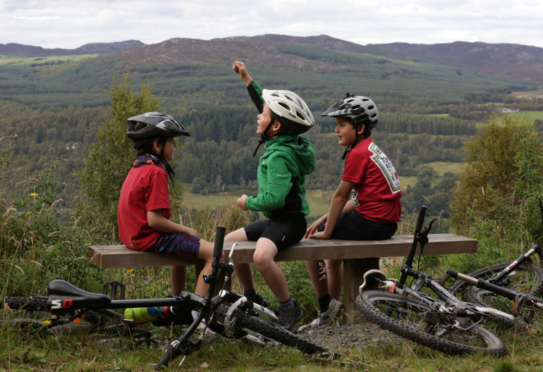  Three young boy cyclists sit on wooden bench high up in Balnain, with views overlooking Loch Ness and hills beyond