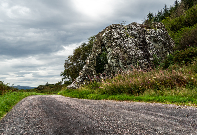  A rocky wall with a hole in the side, next to a lpath with a loch and mountains in the background