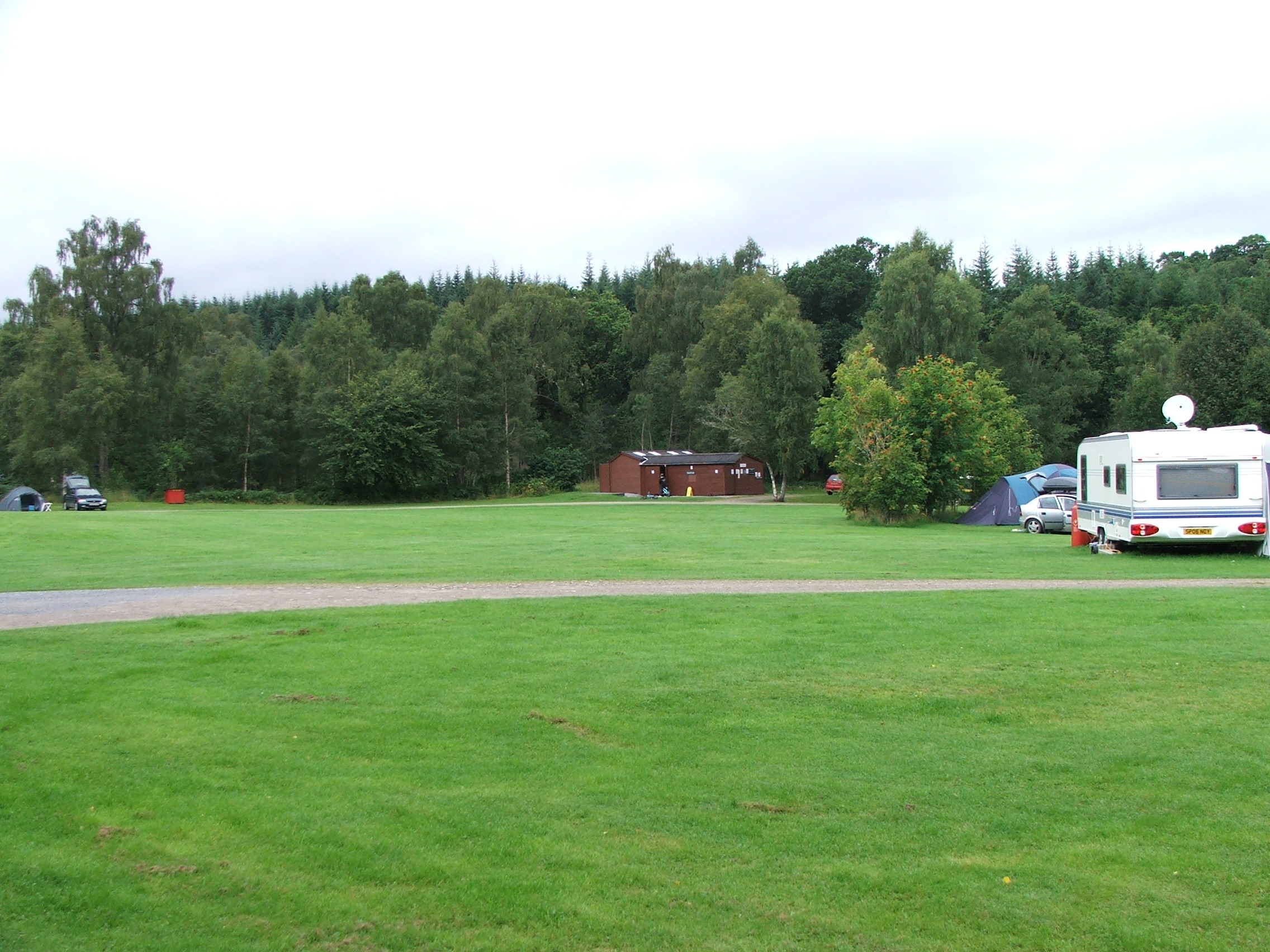 Green grassy field with gravel road through it and trees beyond, with a parked caravan