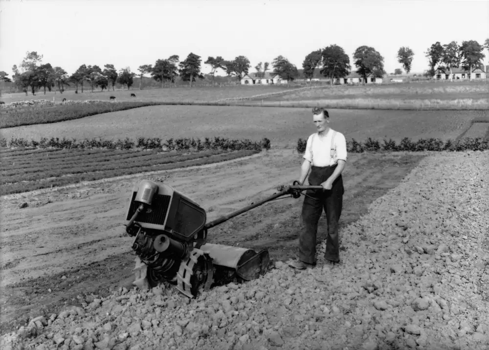 Black & white image of man in shirt moving an old agricultural machine up a plowed field