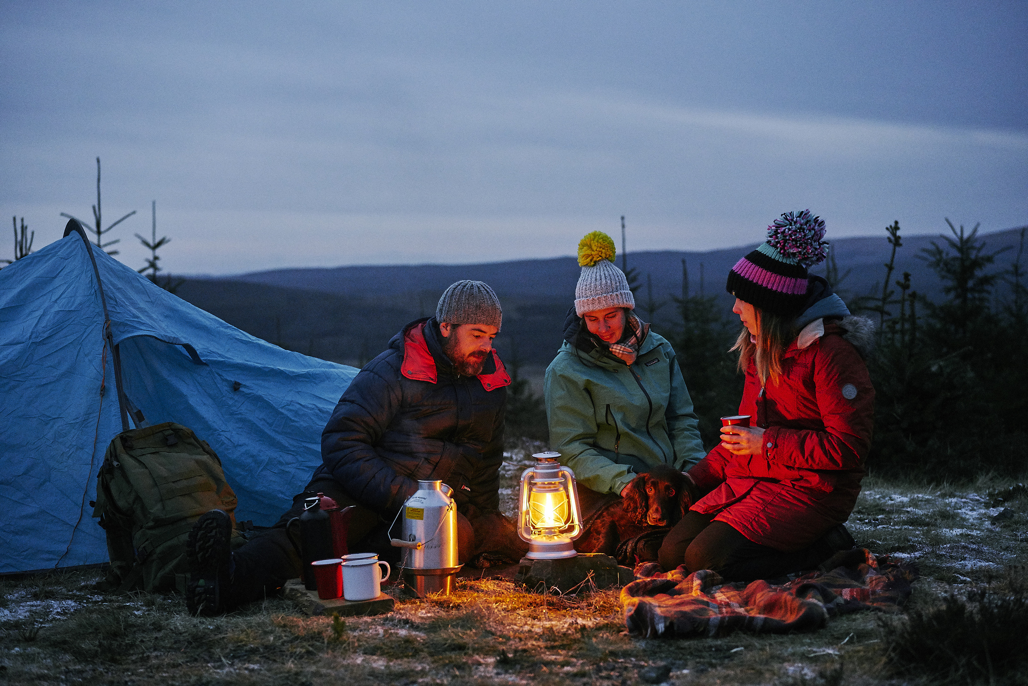 Group of one man and two women campers, dressed in outdoor jackets and hats, sit together outside blue tent at night, with lit hurricane lamp and Kelly camping kettle on ground, and cups of beverage in their hands, and dog lying beside them, Glentrool