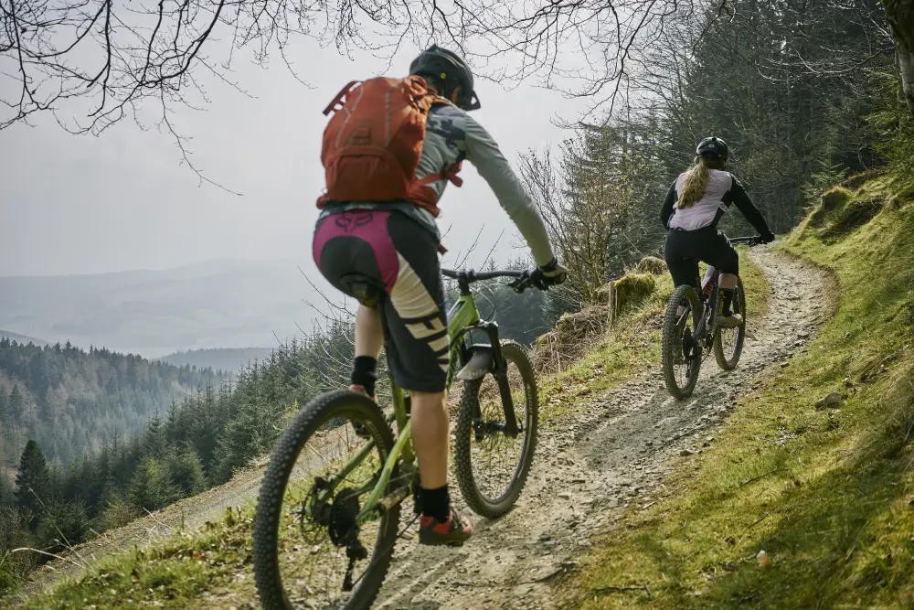 Two people riding mountain bikes up a narrow off road trail with a wide view out and below them across a valley