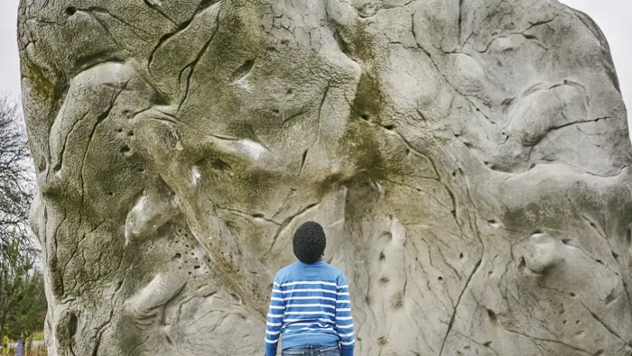  Young boy in jeans and blue and white striped jumper looking up at side of boulder with glimpses of sky at Cuningar Loop Woodland Park, Glasgow