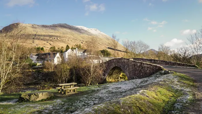 A picnic area and a stone bridge leading to houses with snowy mountains behind