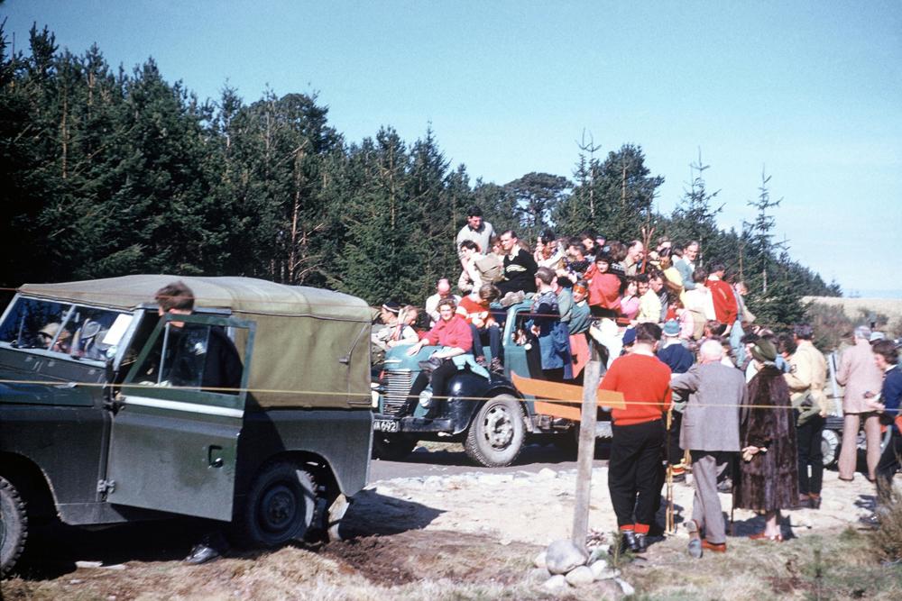 Ski bus that is full of people with people perched on the bonnet and the roof of the cab.
