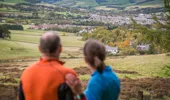 Male runner (in orange top) and female runner (in blue top) on top of hill look over towns in valley, Cademuir Forest, near Peebles